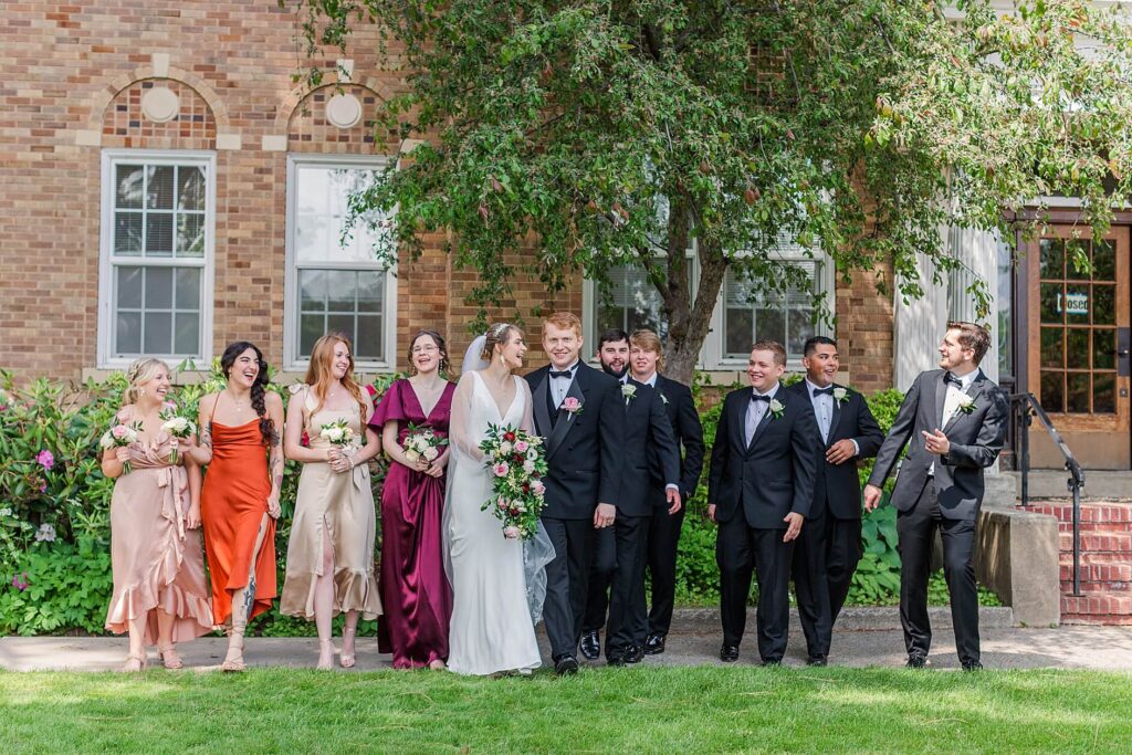 Full Bridal party dressed in fall colors of Burgundy, orange, and cream and guys in black tuxedos walk with the bride and groom in front of the Saint thomas Church in Coeur d'Alene