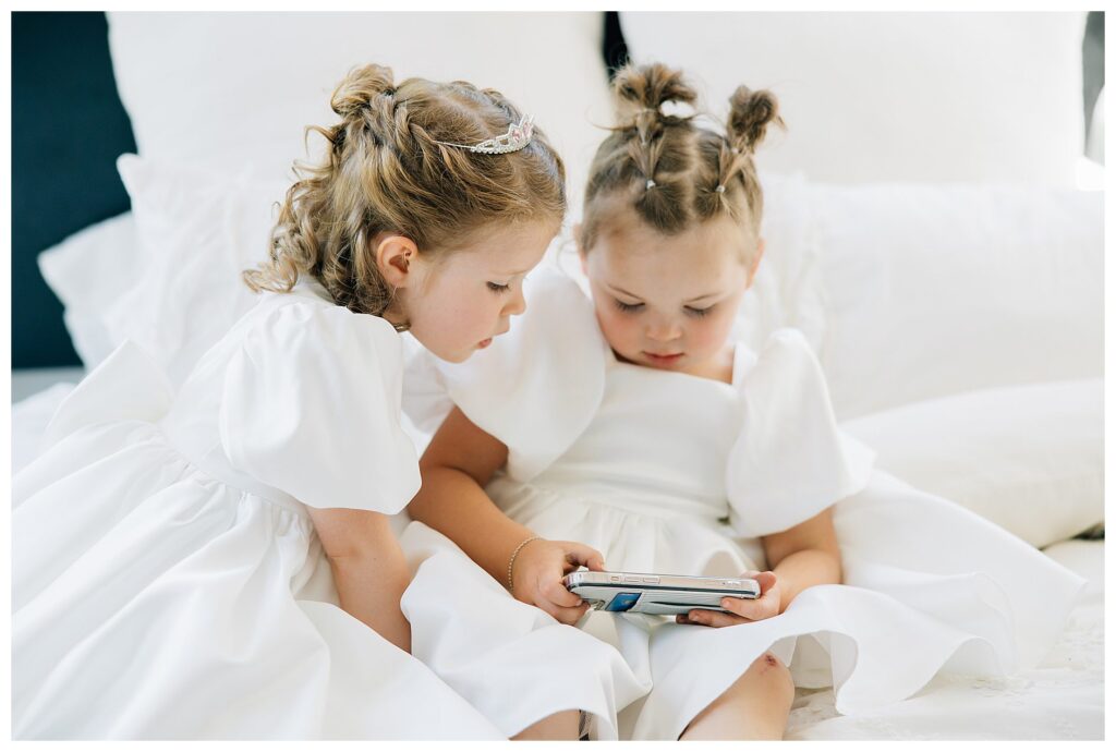Flower girls in white satin flower girl dresses watching a show on the bed while the bride and bridesmaids get ready 
