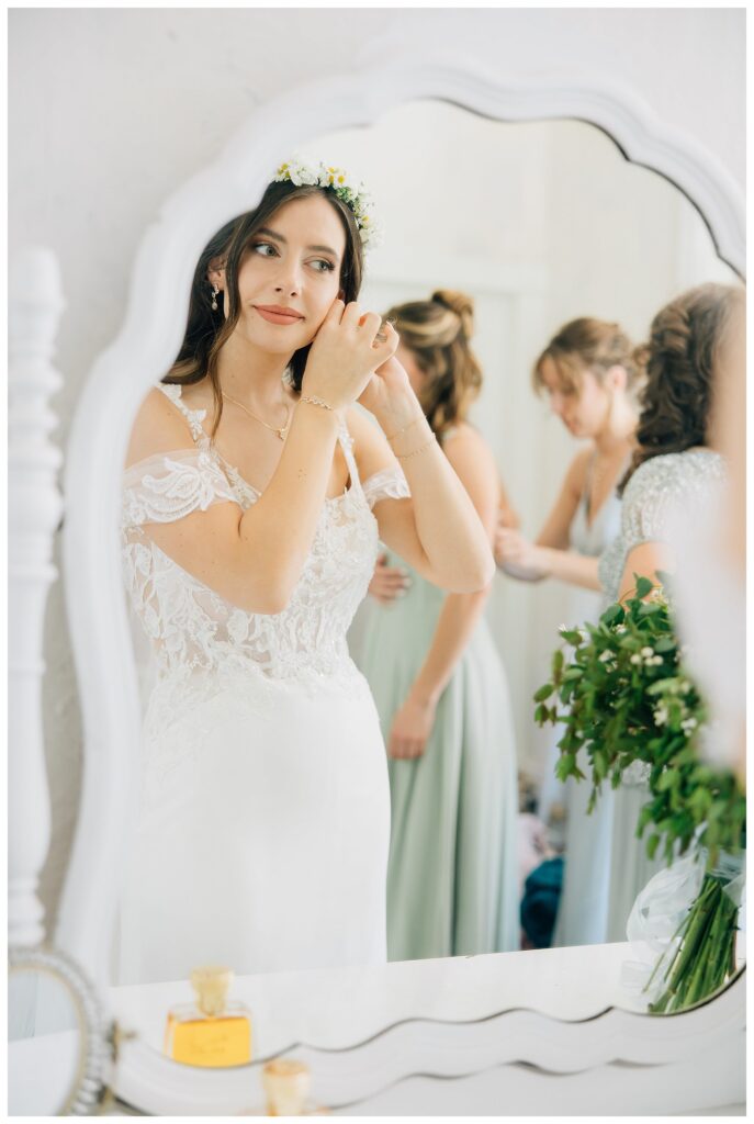 Bride with a daisy flower crown softly placing on her earrings while looking into the mirror