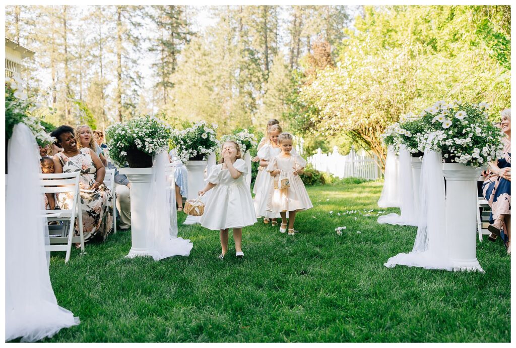 Flower girls in satin dresses walk down the aisle on a summer day at Belle gardens with one girl giggling to herself 