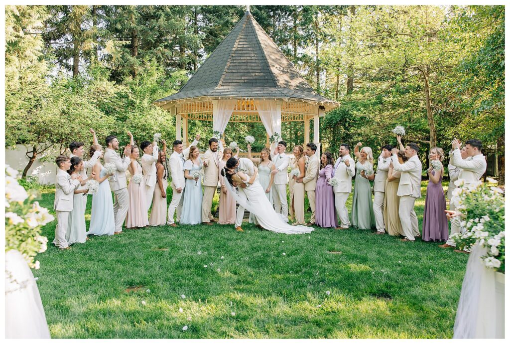 Bridal party in spring pastel wedding colors cheer for the Bride and groom as they kiss in front of a gazebo at Belle gardens