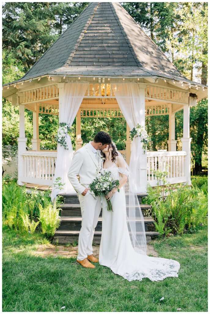 Bride and groom in white and cream share a kiss in front of their Belle Gardens Ceremony gazebo with white and green flowers 